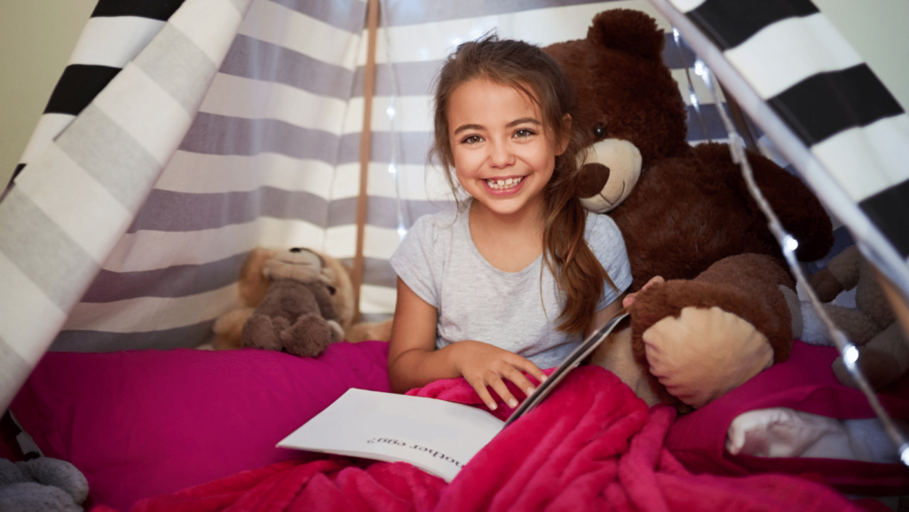 A little girl sitting in a teepee reading a book.