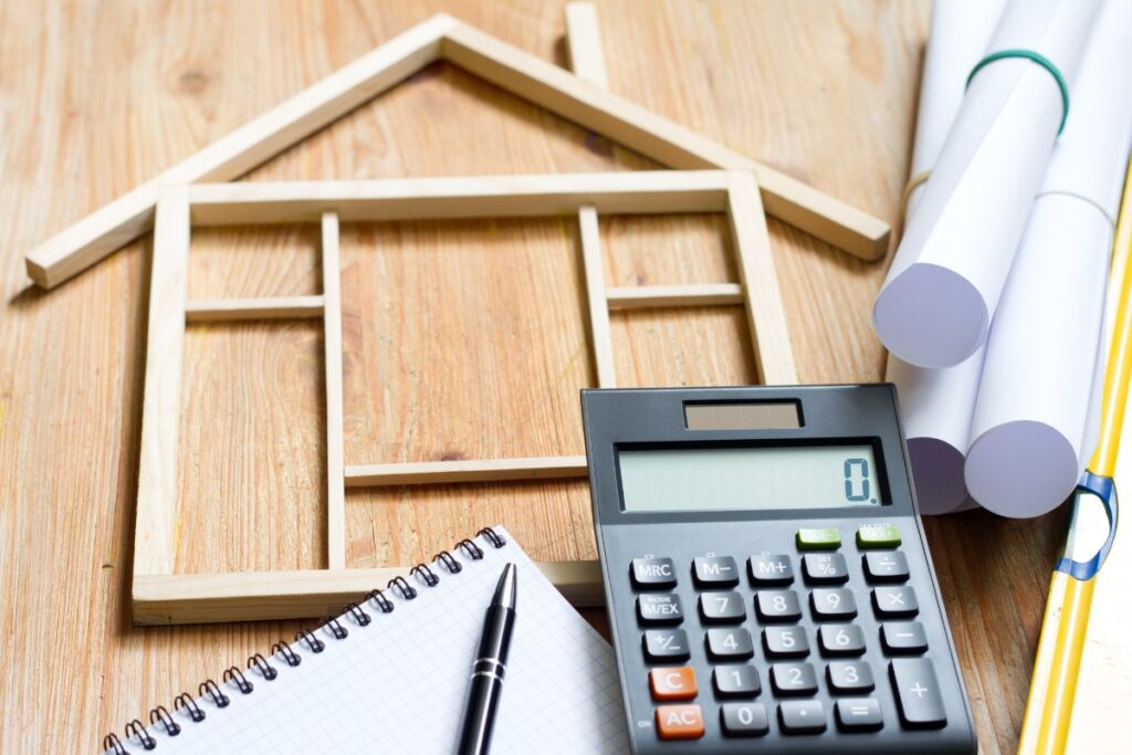 A house plans on a wooden table with a calculator and pen, perfect for creating a remodeling budget.