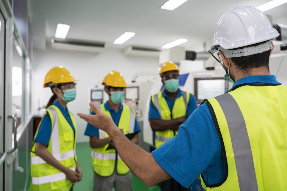 A supervisor in a hard hat and safety vest gestures while addressing a group of attentive industrial workers wearing protective gear about safety standards in construction.