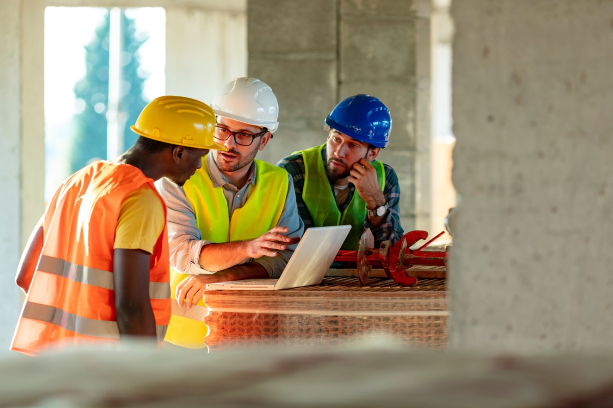 Three construction workers in hard hats discussing safety standards and plans on a laptop at a construction site.