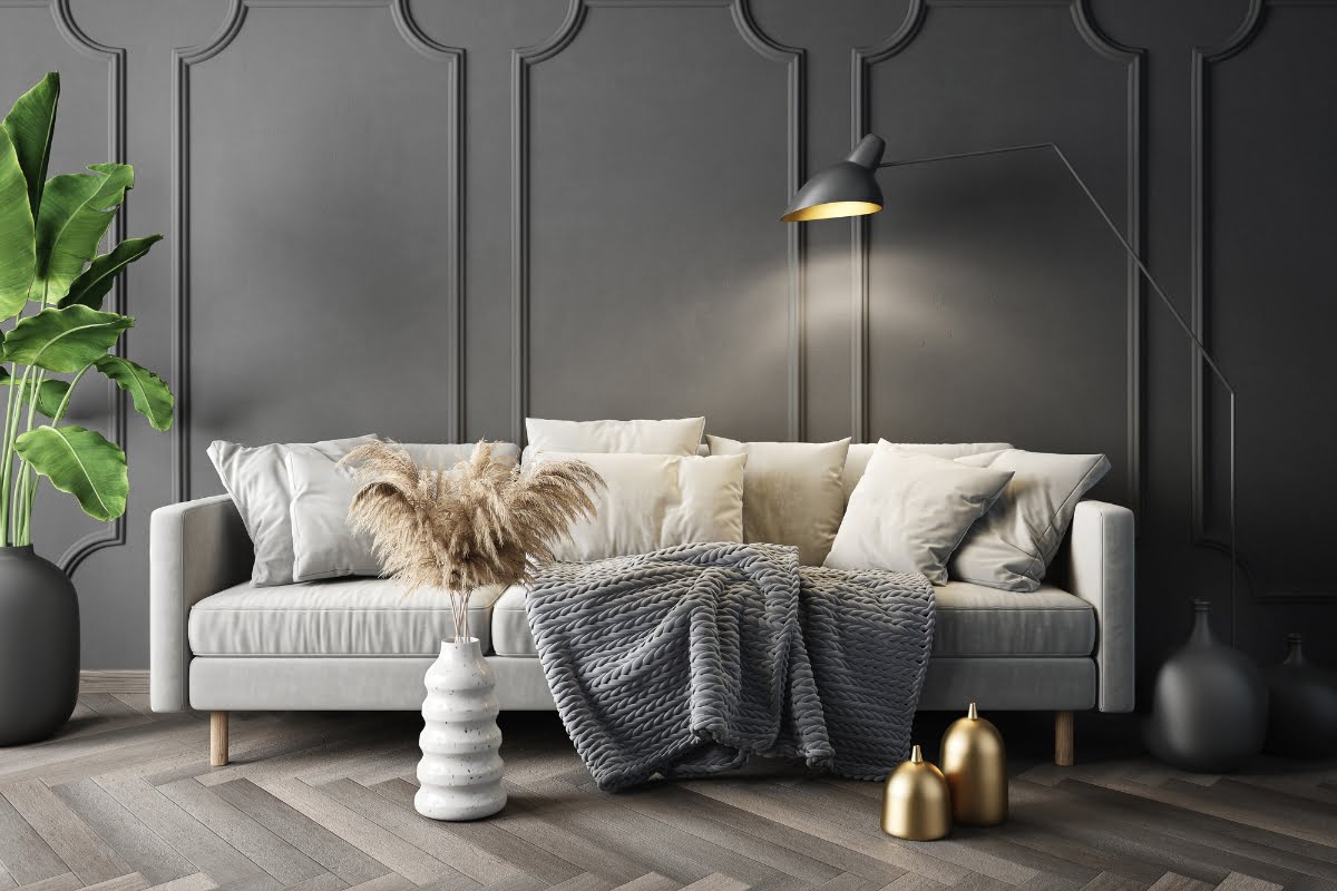 A modern living room features a grey sofa with white and grey pillows, a grey throw, a floor lamp, and decorative vases. Bright green plants add a touch of nature against dark paneled walls, showcasing impeccable home aesthetics and an eye for interior decorating.