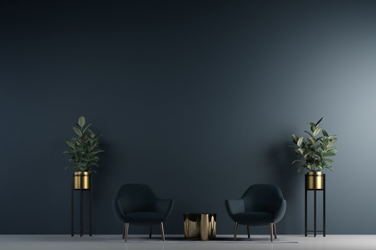 A minimalist room with two dark blue armchairs, a small round table, and two tall potted plants against a dark-colored wall showcases impeccable home aesthetics.