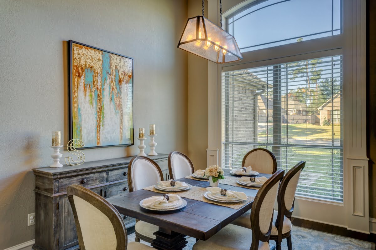 A dining room with a wooden table set for four, beige cushioned chairs, a sideboard with candles and decor, large window with blinds, and wall art that exemplifies refined home aesthetics. Perfect for those who appreciate interior decorating at its finest.