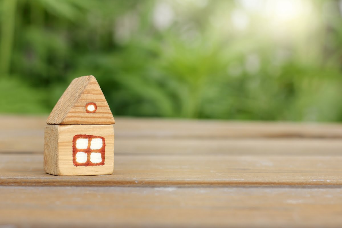 A small wooden house model on a wooden surface representing real estate market trends, with a blurred green garden background lit by sunlight.