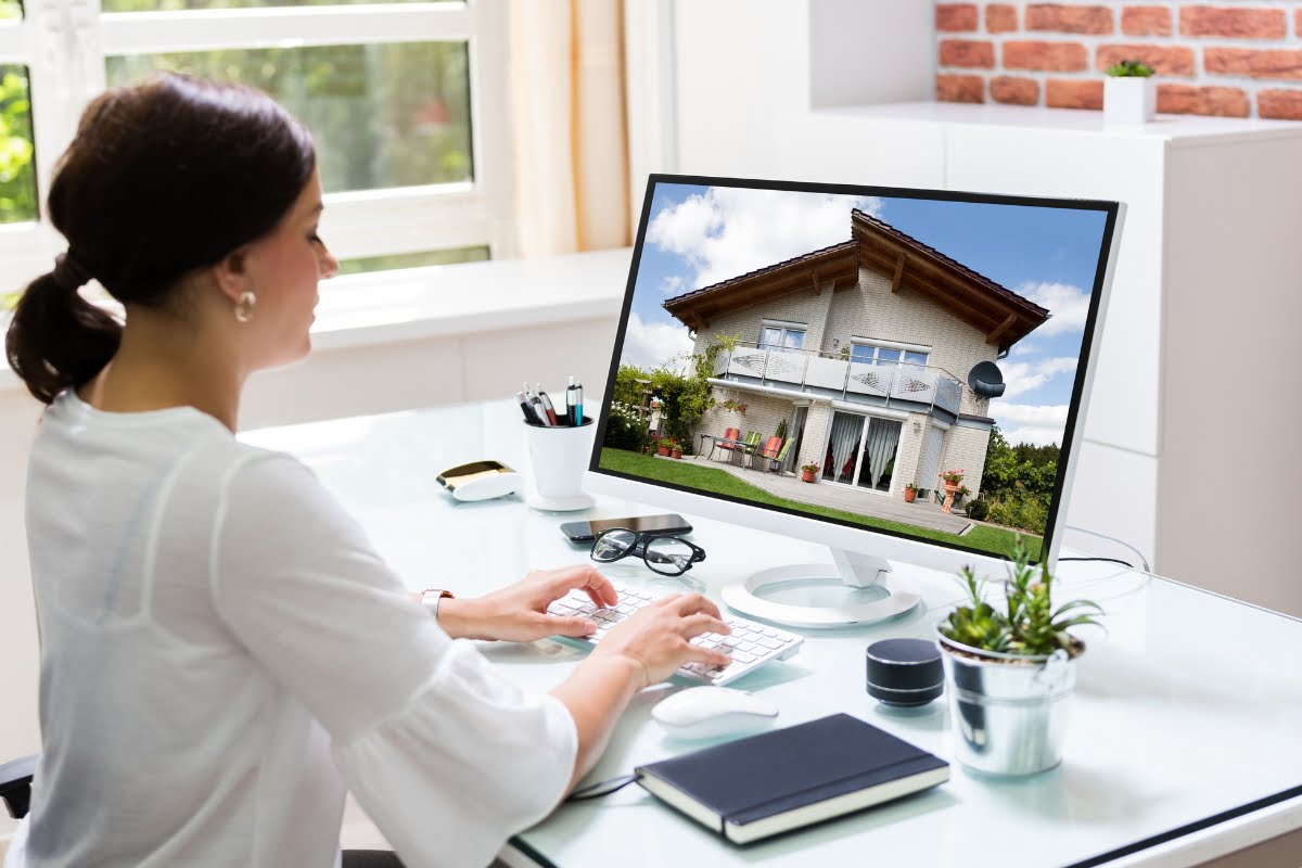 A woman in a white shirt analyzes real estate market trends on a computer displaying a house image in a bright office setting.