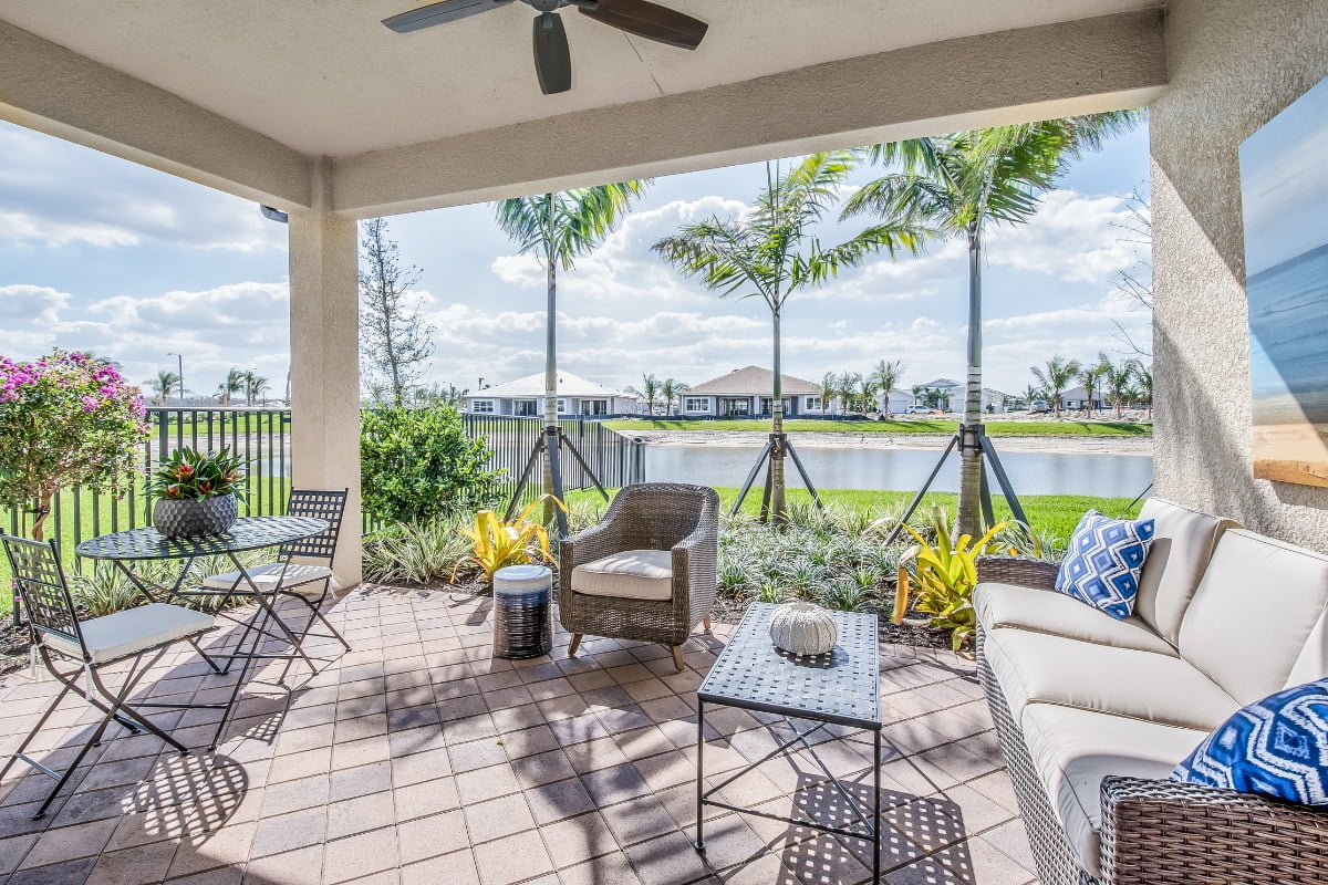 Patio with seating area overlooking a lake, showcasing real estate market trends, featuring a ceiling fan, tables, chairs, and lush plants under a clear blue sky.