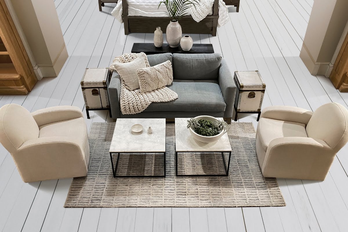 A modern living room with a gray sofa, two beige armchairs, and two metal-framed coffee tables on a beige rug. Decor includes pillows, plants, and minimalist items influenced by real estate