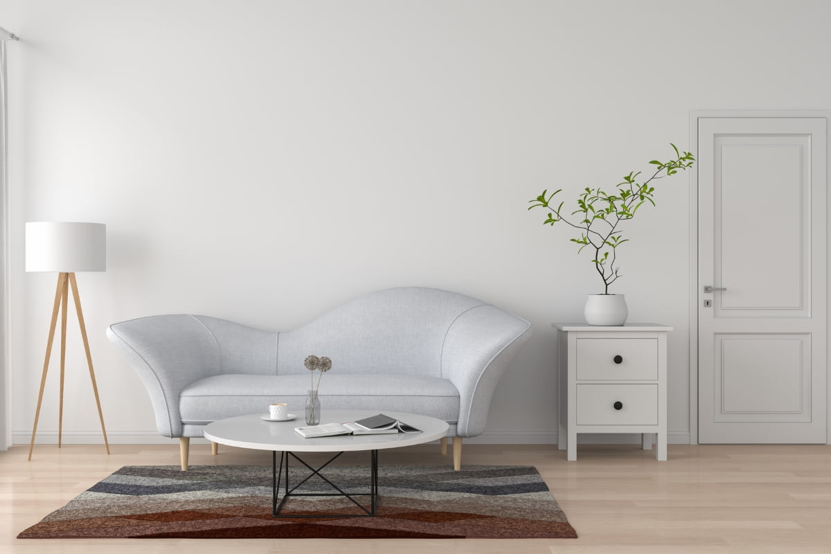 A minimalist living room with a gray sofa, white round table, lamp, and a plant next to a small cabinet by a closed door, reflecting current real estate market trends.