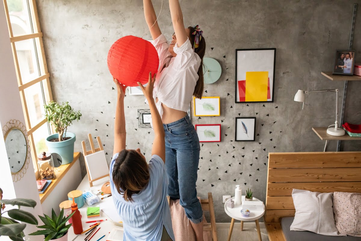 Two people are decorating a room, with one person standing on a ladder hanging a red paper lantern while the other offers diy home improvement tips. The room has framed pictures on the wall and various decor items around.