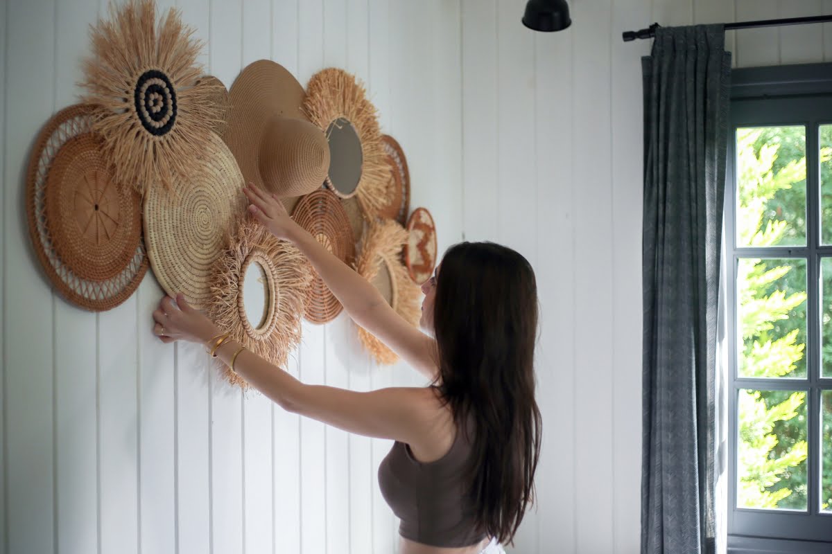 A woman with long hair decorates a white wooden wall with woven baskets of various sizes and shapes, some of which have mirrors. There is a window with gray curtains beside her, adding texture and warmth to the space. This scene exemplifies diy home improvement tips for creating personalized decor.