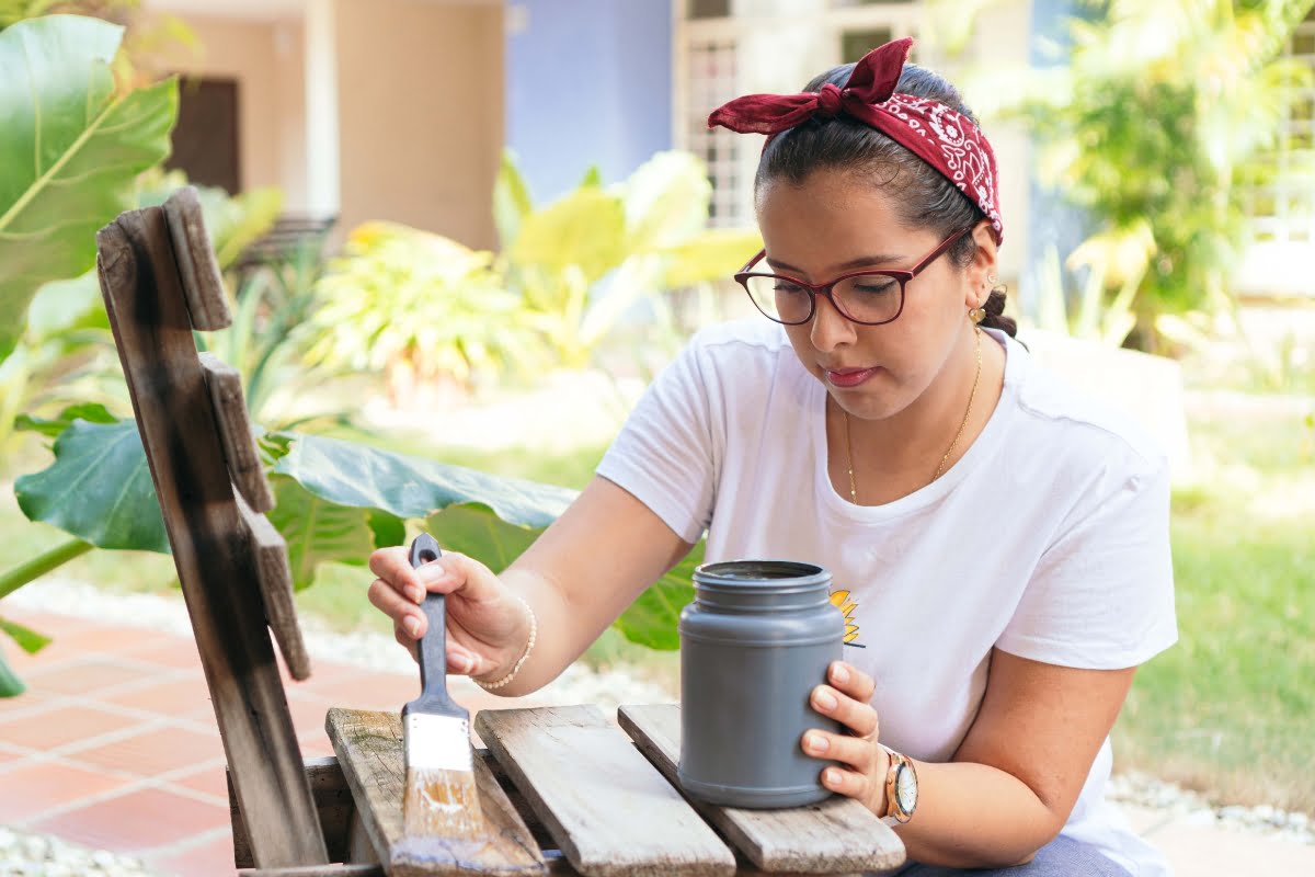 A person with glasses and a red bandana is painting a wooden chair outdoors using a brush and a can of paint, embodying some effective DIY home improvement tips.