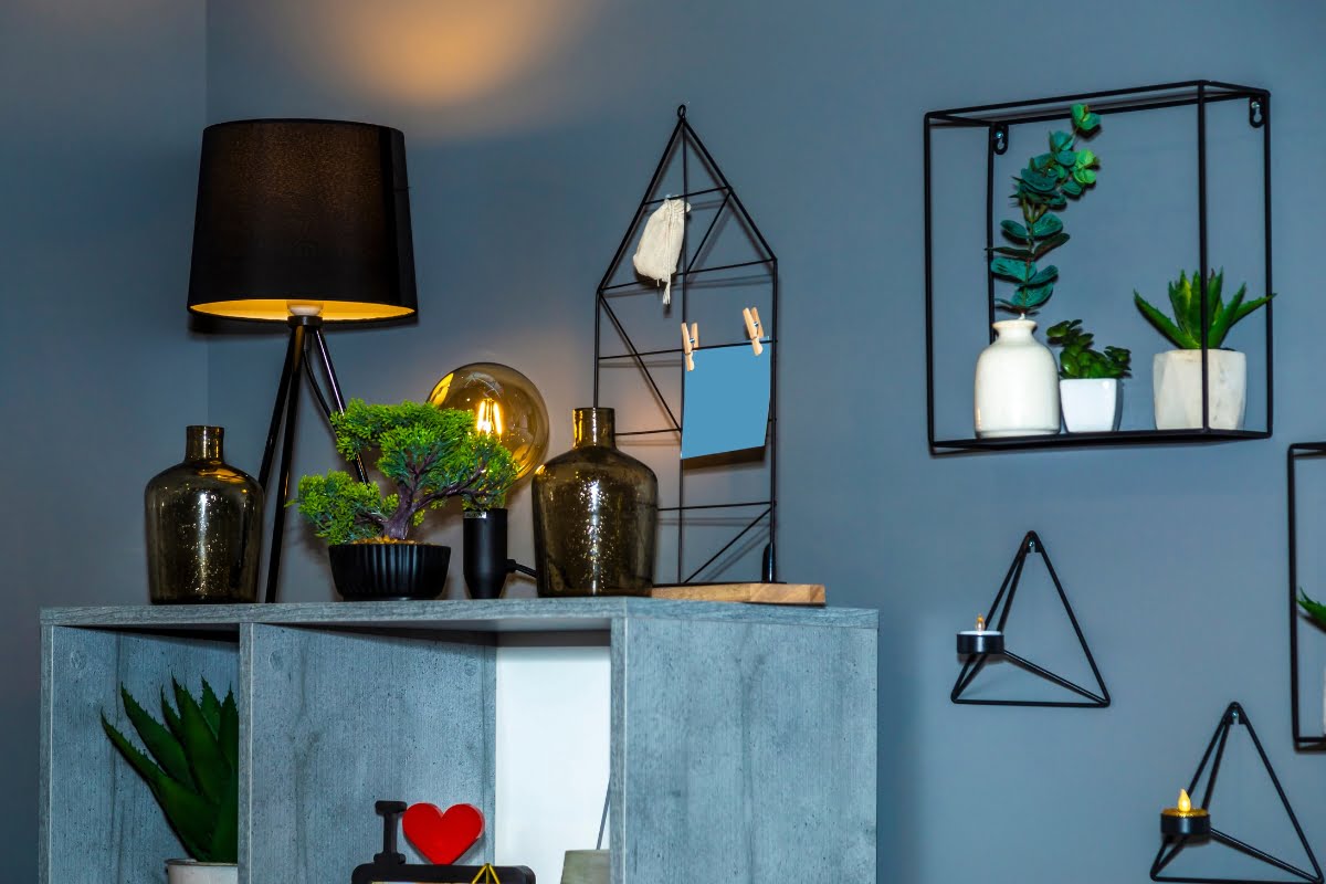 A modern living room corner featuring a gray shelf adorned with plants, vases, and decorative items. Above, various geometric wall shelves hold more plants and ornaments. A black lamp is on the shelf, creating a perfect setting to showcase your diy home improvement tips.