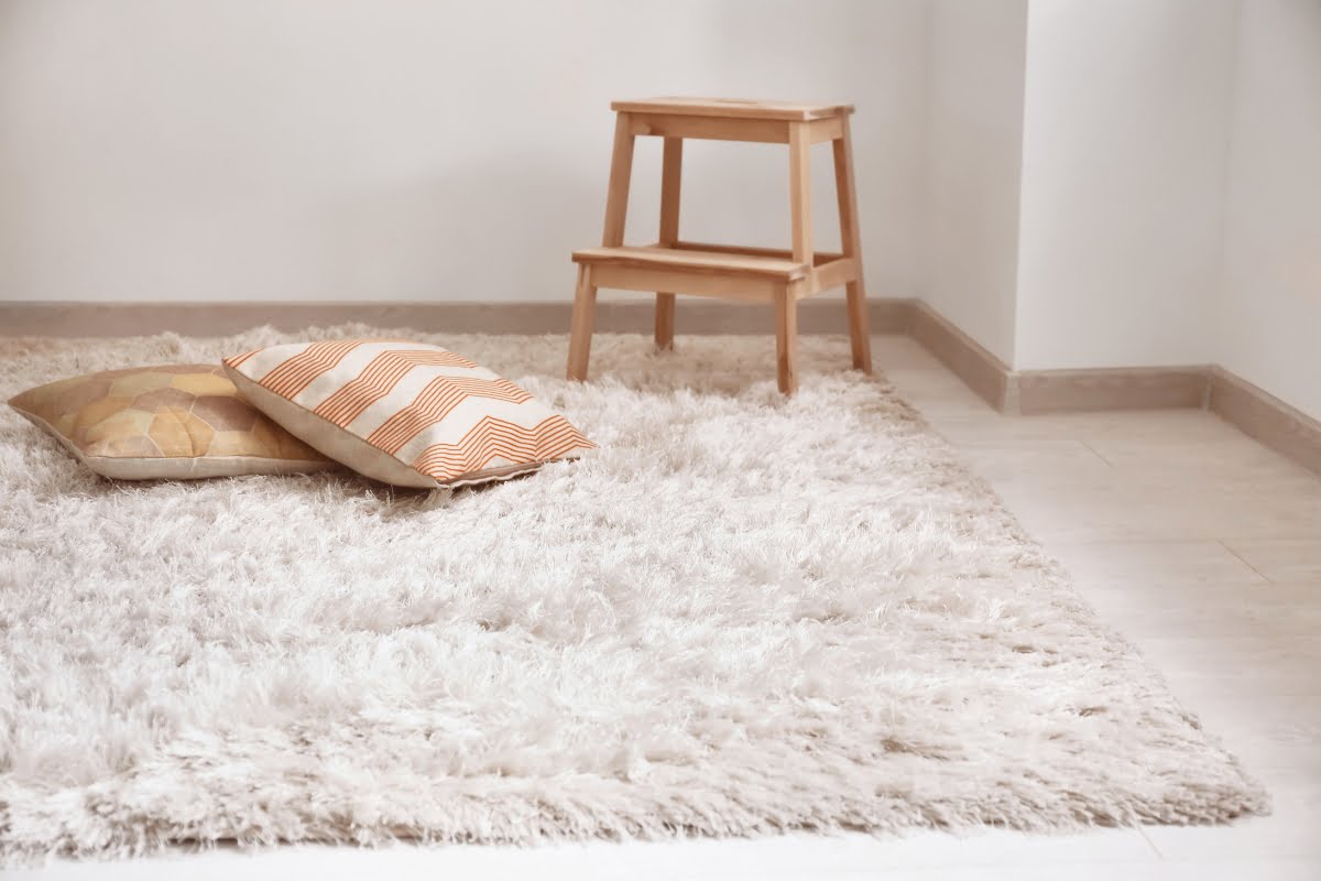 A light-colored fluffy rug with two patterned throw pillows is on the floor. A wooden step stool stands nearby in a minimally decorated room, perfect for those seeking diy home improvement tips to create a cozy yet simple space.