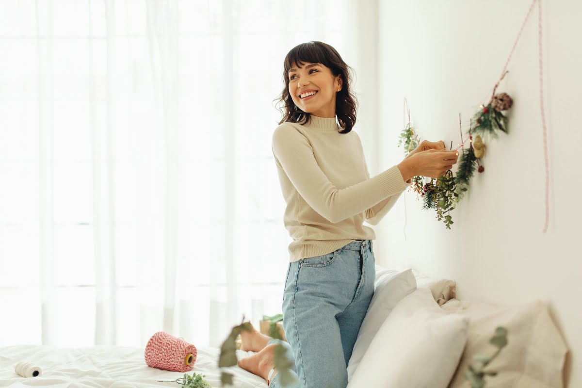 A woman stands on a bed, decorating a wall with hanging greenery and flowers. She is smiling and wearing a beige sweater and blue jeans, embodying DIY home improvement tips. The room is bright with natural light from the window, making her project glow.