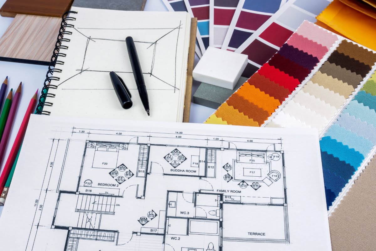 A desk with architectural floor plans, color swatches, fabric samples, a notebook, pens, pencils, a color palette, and some DIY home improvement tips.