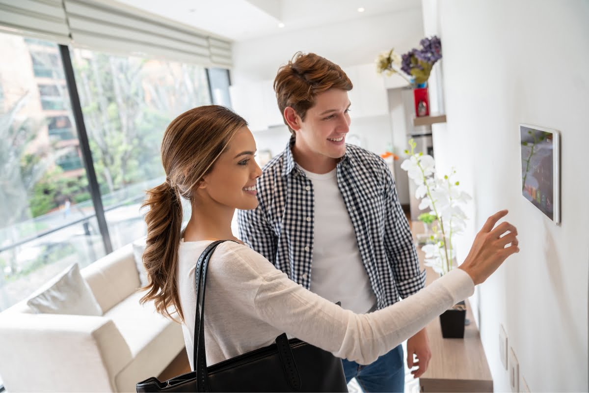 A woman and man stand in a modern living room, interacting with a touchscreen control panel mounted on the wall, seamlessly integrating smart home technology into their daily routine.