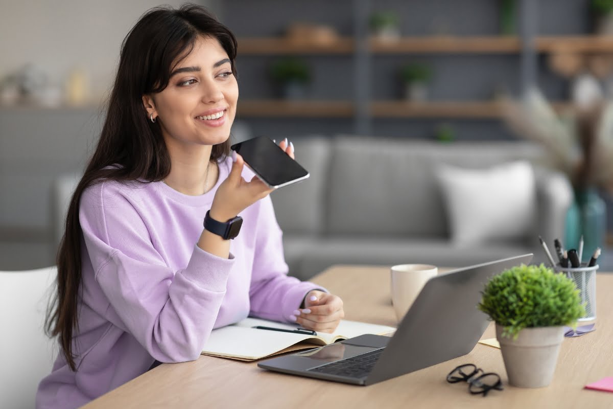 A woman in a purple sweater is sitting at a desk with a laptop, notebook, and mug, speaking into a smartphone. A pot of green plants is on the desk. Shelves with decor and smart home technology are in the background.