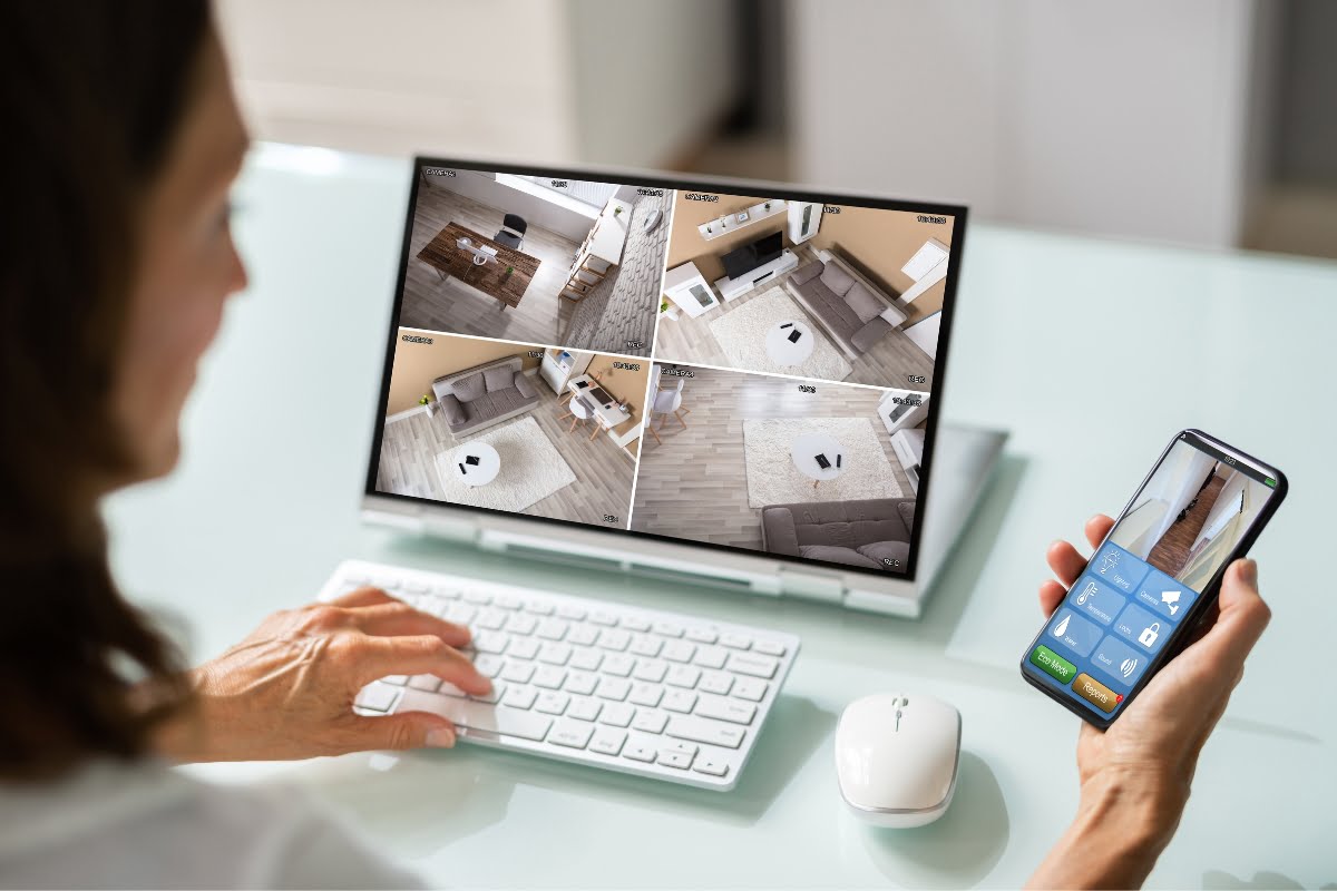 A person monitors multiple home security feeds on a computer screen and controls settings using a smartphone, showcasing the convenience of smart home technology in a modern indoor setting.