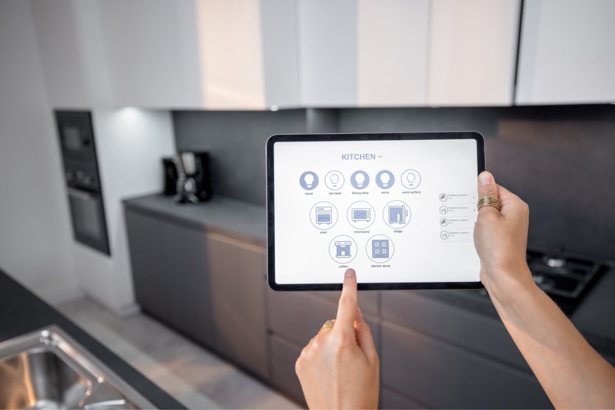 A person uses a tablet to control smart kitchen devices, including lights, oven, coffee machine, and music system. The tablet displays various control icons in a sleek, modern kitchen setting—showcasing the convenience of Smart Home Technology.