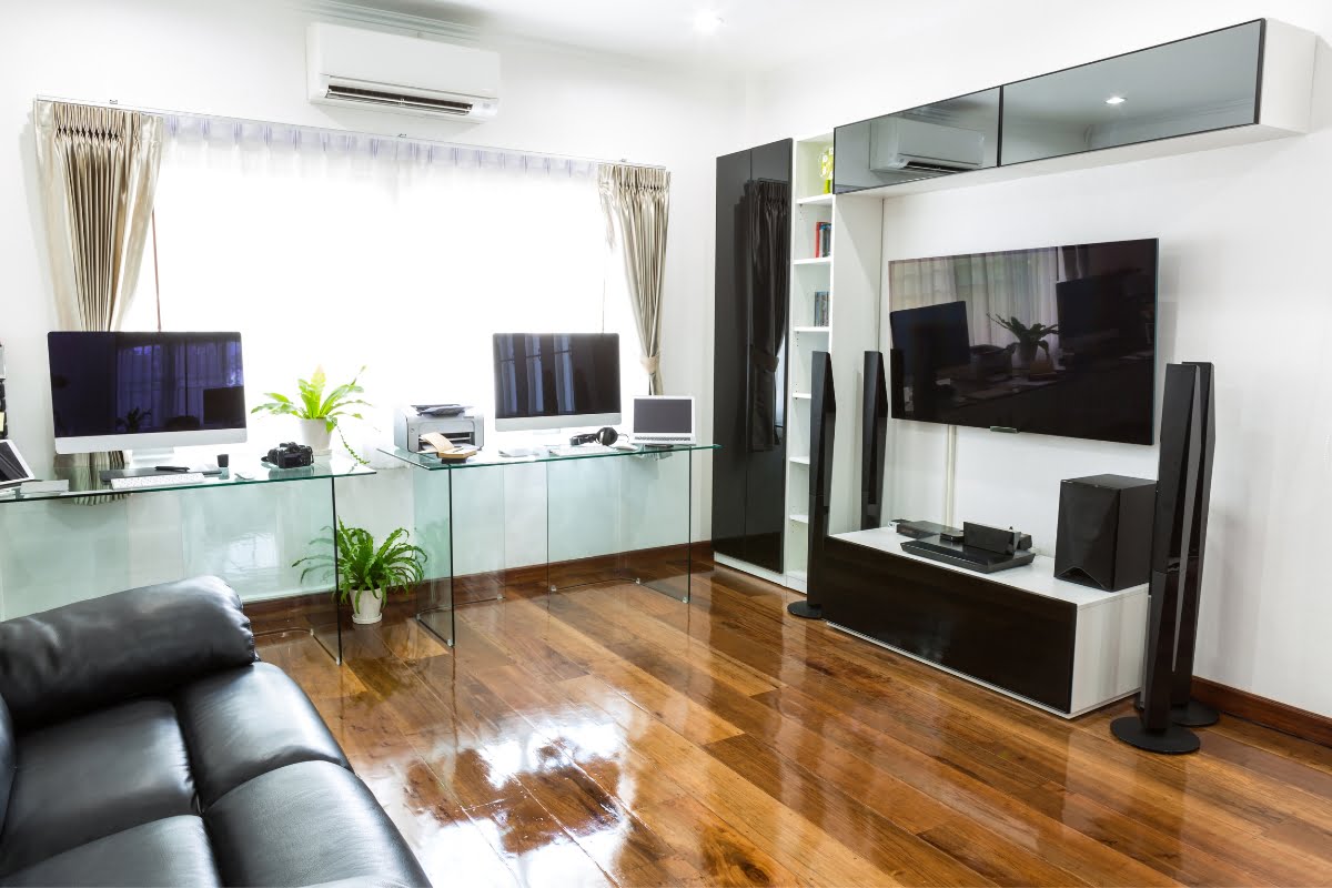 A modern home office with smart home technology features two computer setups on a glass desk, a large TV with sound system, minimalist black and white furniture, a leather sofa, and wooden flooring.