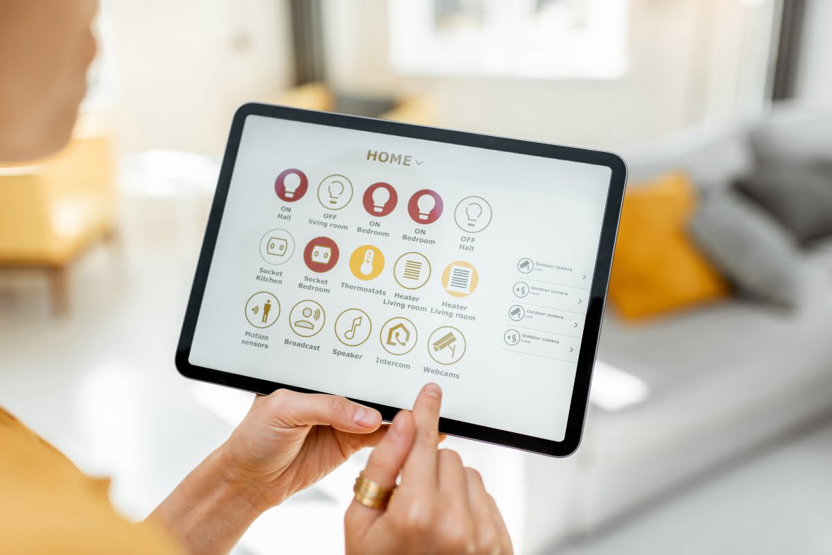 A person uses a tablet displaying a home automation app interface, with icons for controlling various household devices and settings, showcasing the convenience of smart home technology.