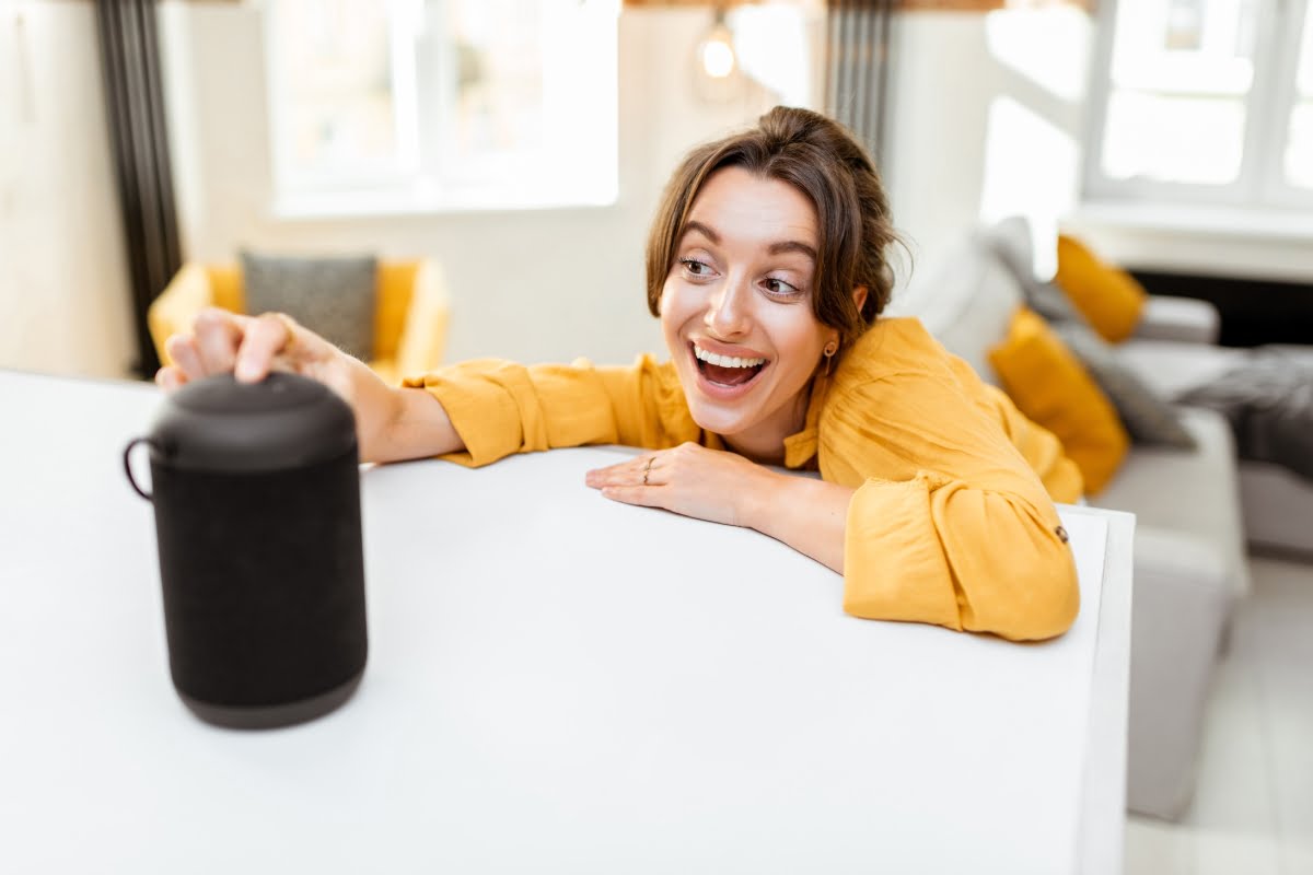 Person in a yellow shirt smiling and reaching for a black portable speaker on a white surface in a brightly lit room with yellow and gray decor, showcasing the integration of Smart Home Technology.