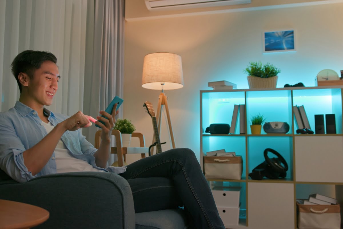 A man sitting on a couch in a warmly lit living room uses his smartphone. The cozy space features shelves and a lamp in the background, seamlessly integrating smart home technology.