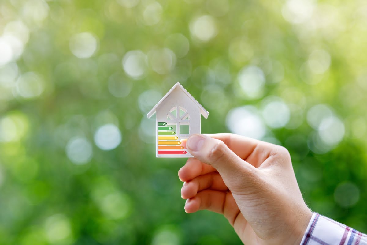 A hand holds a small house-shaped cutout with an energy efficiency rating chart against a blurred green background, highlighting the importance of energy efficiency in homes.