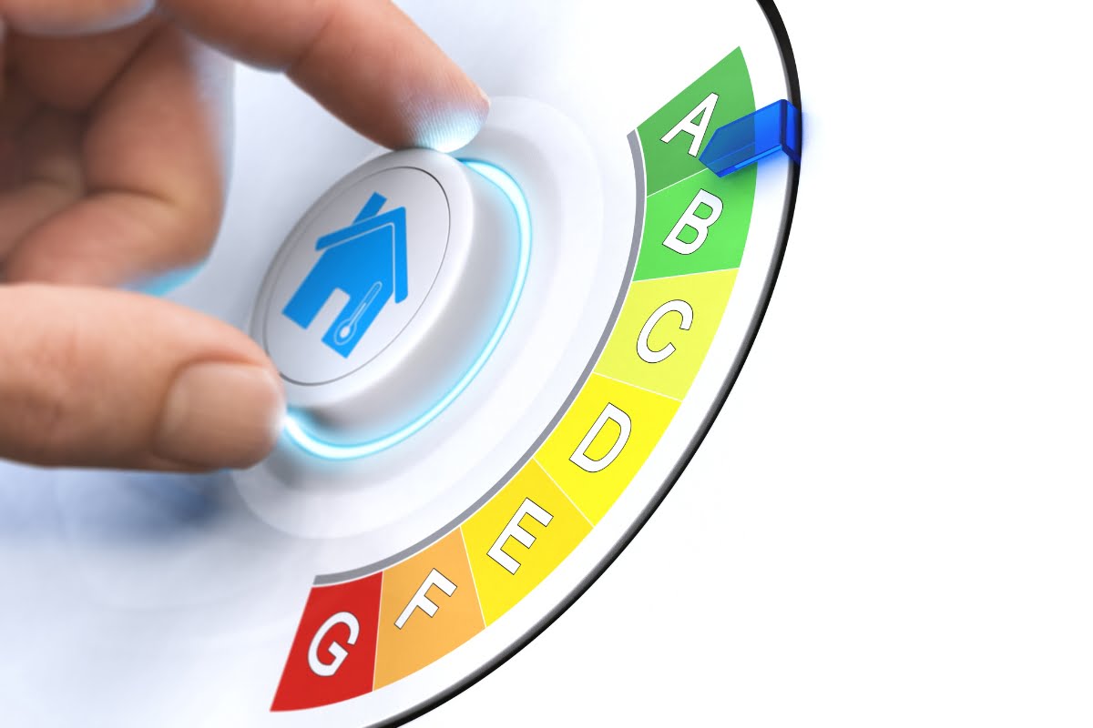 A hand turns a dial with an energy efficiency indicator, moving the pointer from lower (red) to higher (green) letters. The dial, emphasizing energy efficiency in homes, features letters from G to A with a blue house symbol at the center.