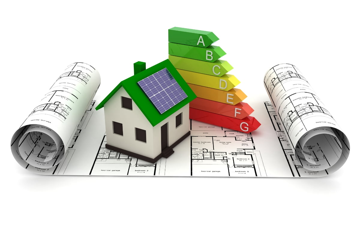 A small house model with a solar panel on the roof stands on architectural blueprints, illustrating Energy Efficiency in Homes, next to a vertical colored energy efficiency scale from A (green) to G (red).