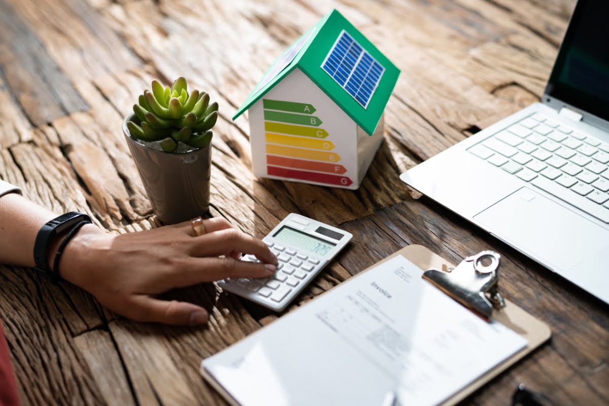 A hand using a calculator next to a model house with an energy efficiency chart, emphasizing Energy Efficiency in Homes, a potted plant, a clipboard with papers, and a laptop on a wooden table.