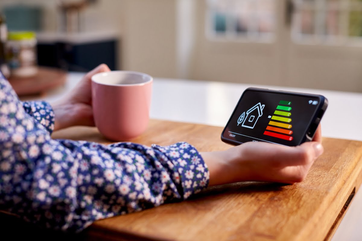 Person holding a pink mug and a smartphone displaying a home energy usage app with a colored bar chart on a wooden table, showcasing the importance of energy efficiency in homes.