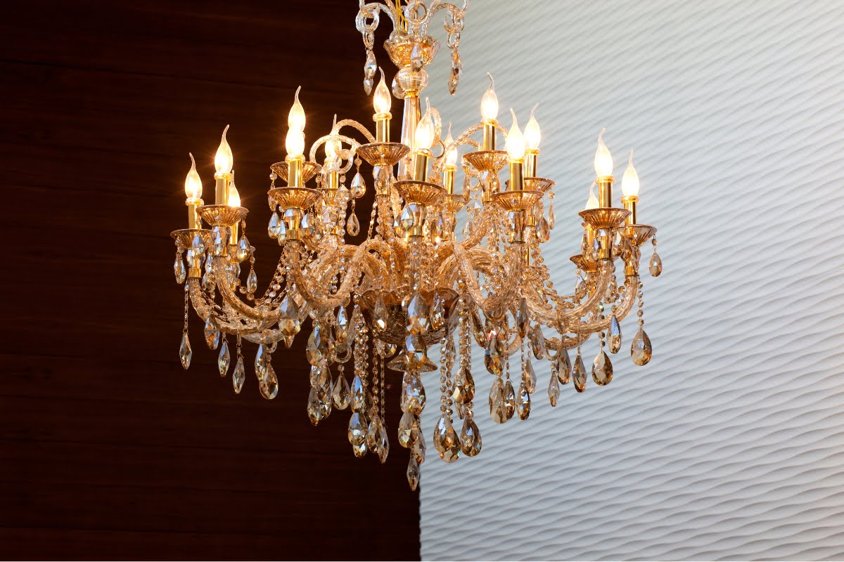 A large ornate crystal chandelier with multiple lit candle-style bulbs hangs against a two-toned brown and white textured background, showcasing exquisite furniture design.