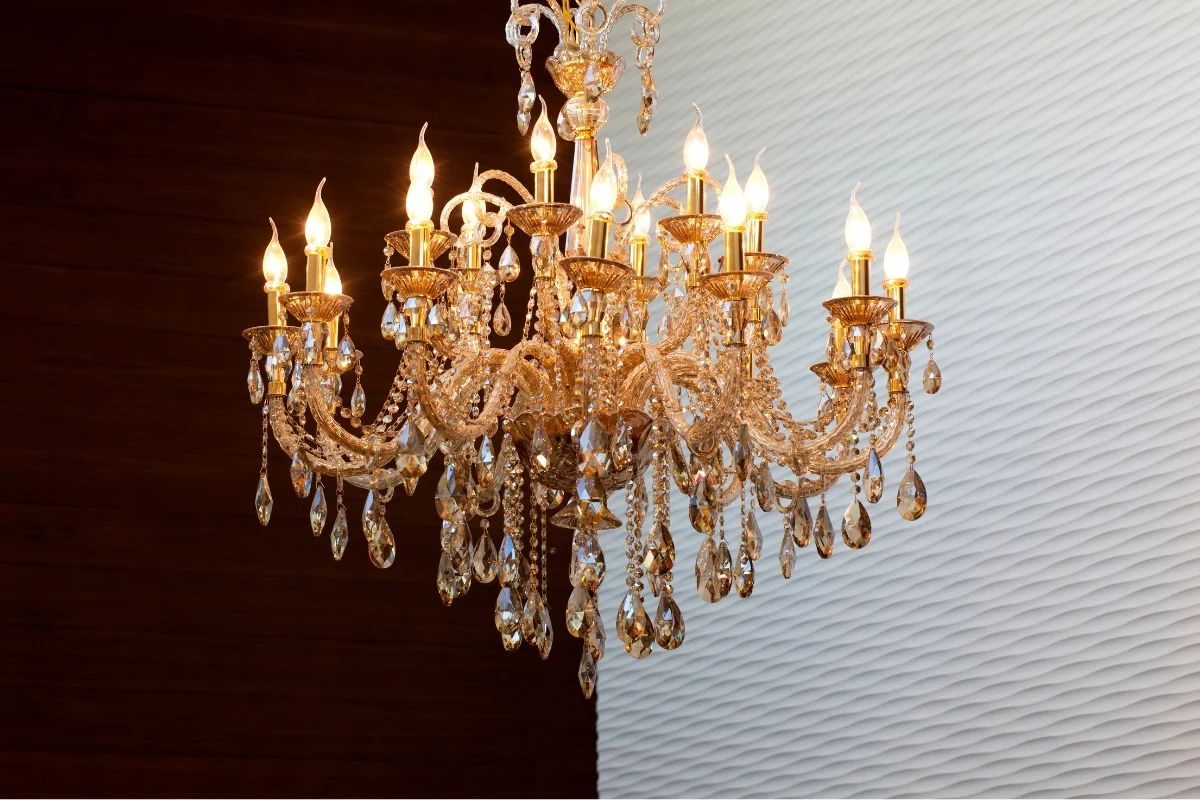 A large ornate crystal chandelier with multiple lit candle-style bulbs hangs against a two-toned brown and white textured background, showcasing exquisite furniture design.