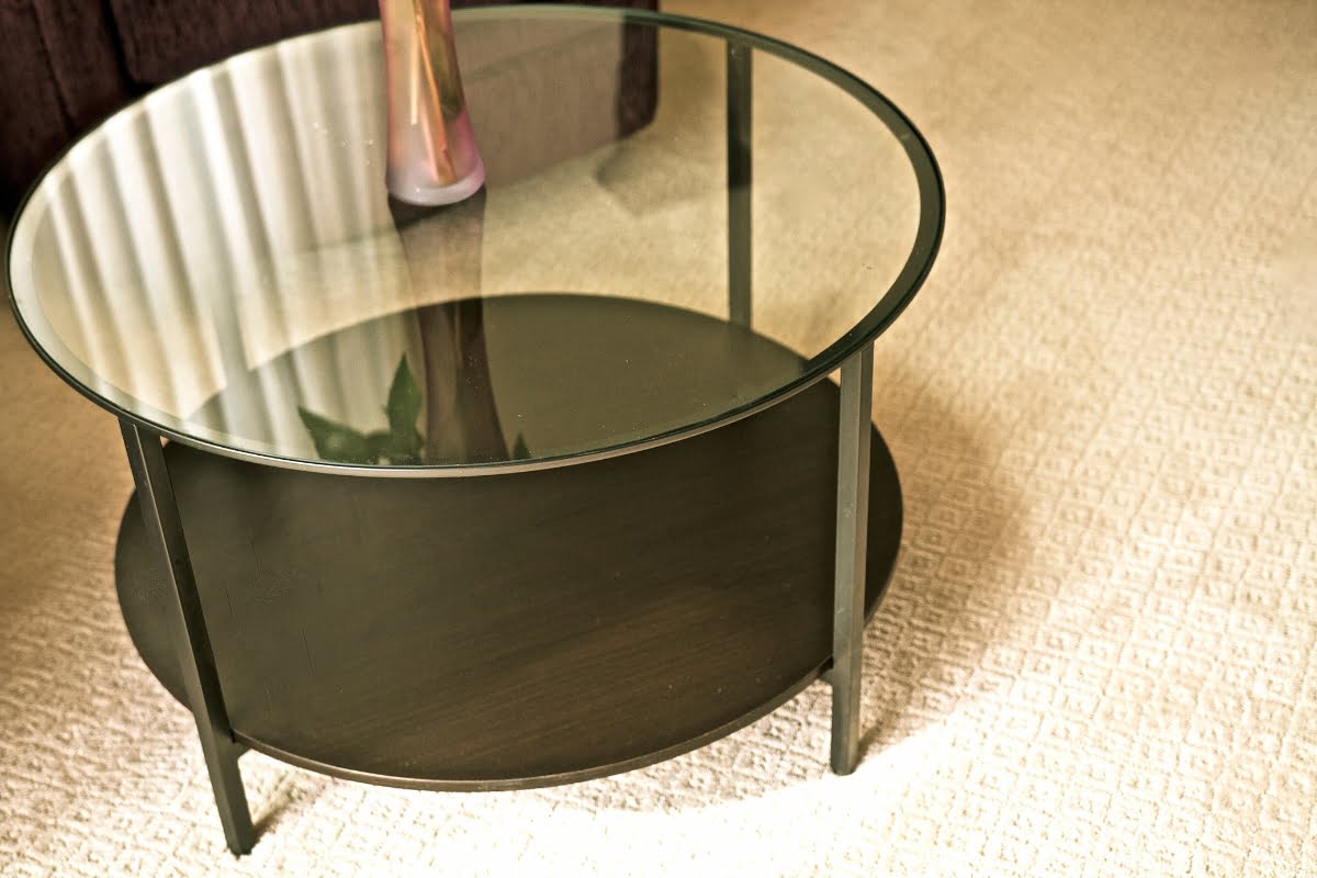 A round glass-top coffee table with a black metal frame and a lower black shelf, showcasing sleek furniture design, is placed on a beige carpet in the living room.