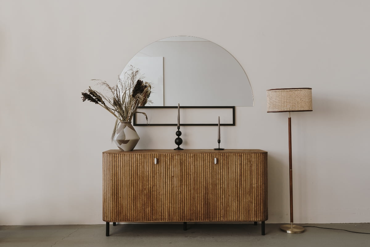 A wooden console with dried flowers in a vase, two candlesticks, and a semi-circular mirror above; next to it, a standing lamp with a woven shade against a plain white wall exemplifies exquisite furniture design.