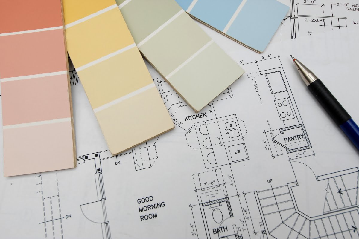 A collection of paint swatches lies on top of a house floor plan with a pen beside them, perfectly complementing the intricate furniture design sketches that decorate the margins.