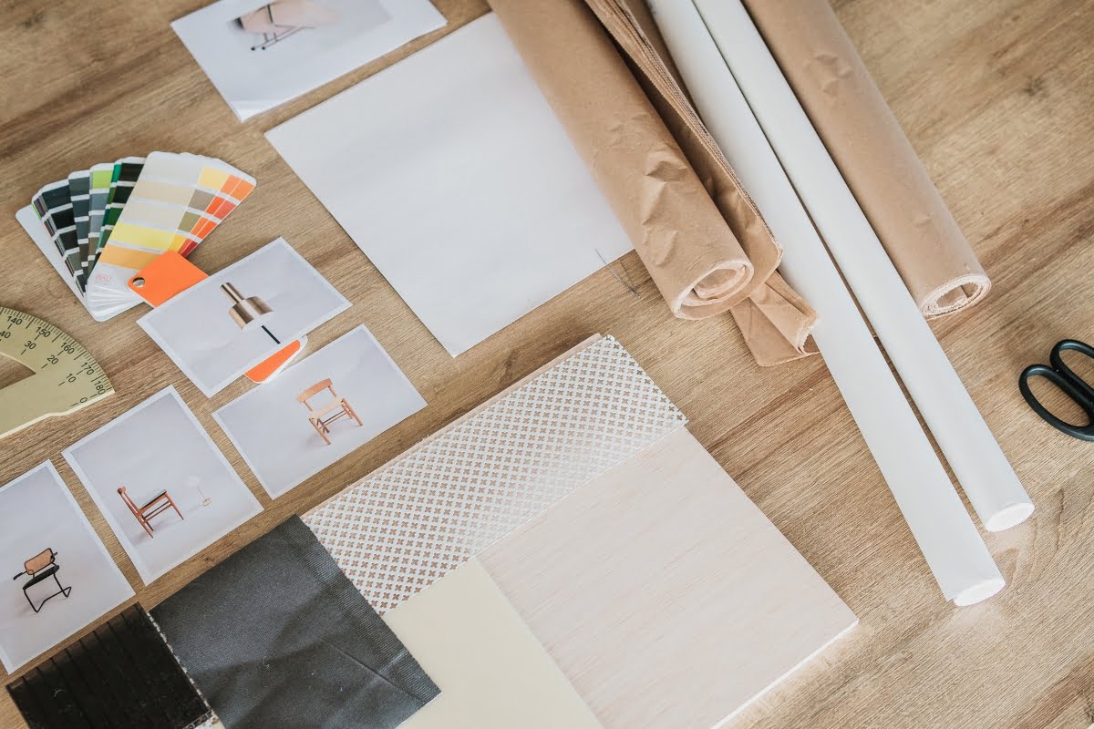 Assorted design materials including fabric swatches, color samples, printed images of furniture design, rolled paper, white boards, and scissors on a wooden table.