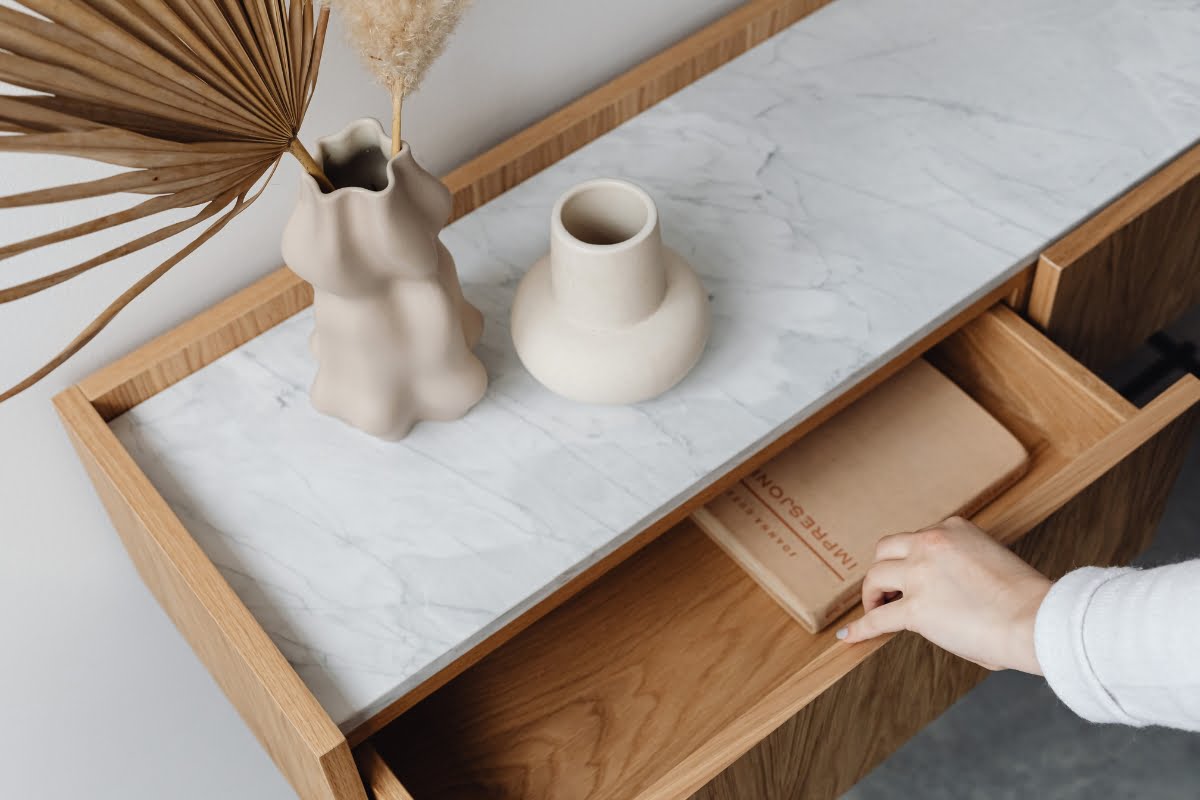 A hand pulls open a wooden drawer with a book inside, revealing a marble-topped table showcasing furniture design with a beige vase and a dried plant arrangement.