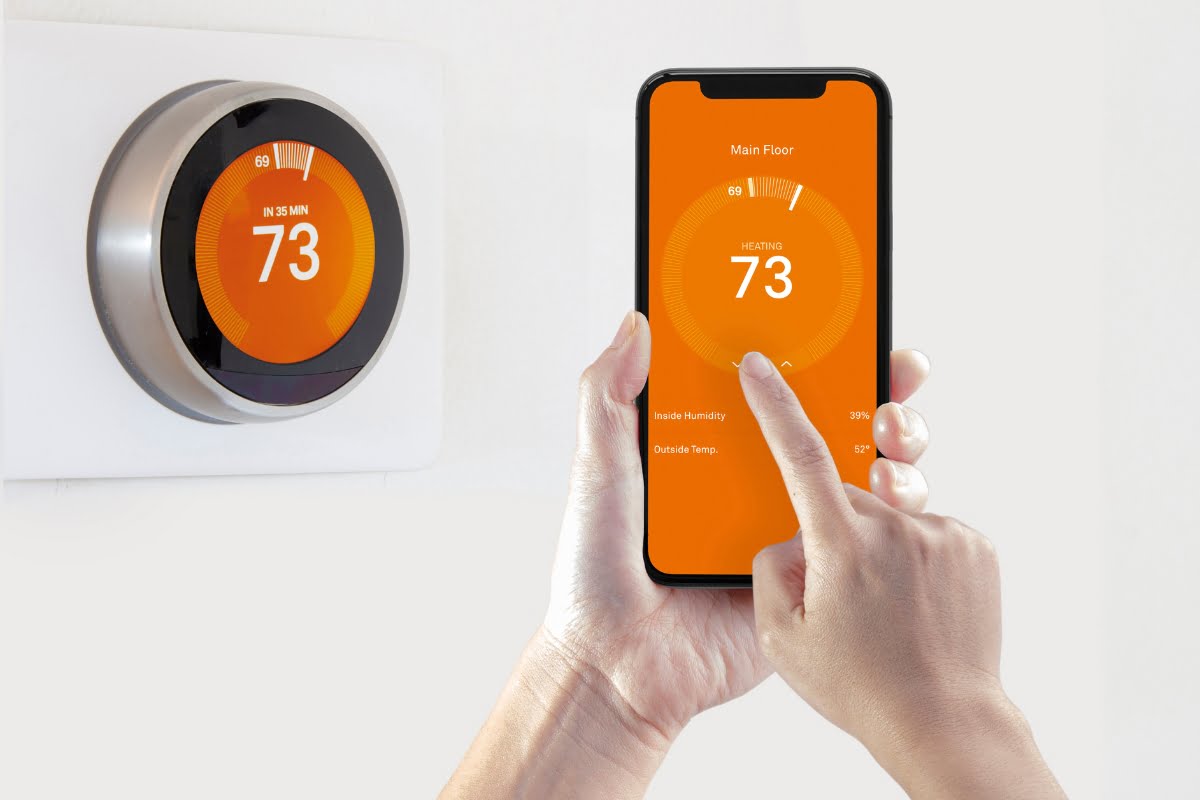 A person adjusts a smart thermostat using a smartphone app, seamlessly integrating with their home automation system. Both the thermostat and the app display the temperature as 73 degrees.