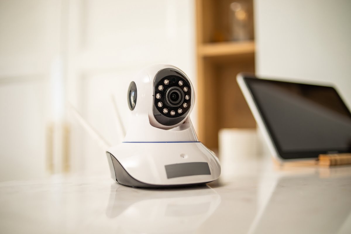 A white security camera, integral to home automation systems, is positioned on a countertop in front of a blurred background that includes a tablet and some shelves.