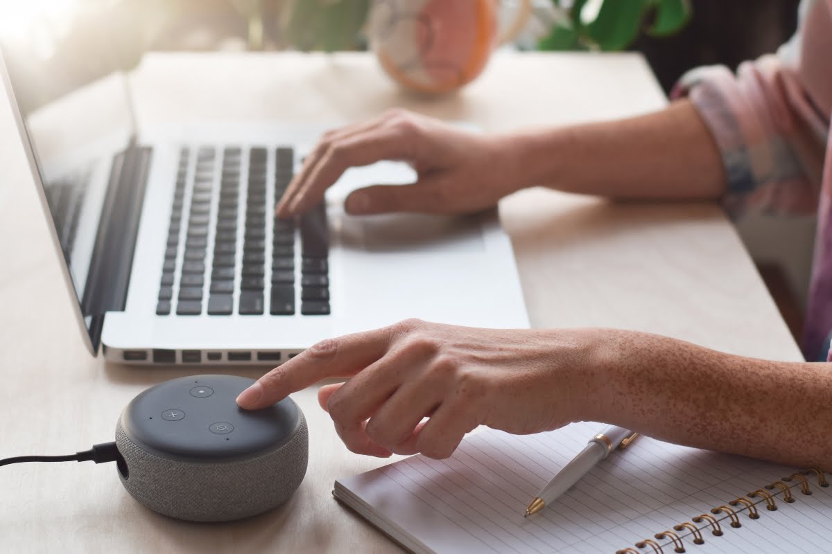 Person typing on a laptop with one hand and using a voice assistant device with the other; notebook and pen are on the table. It's clear they're seamlessly integrating home automation systems into their workflow.