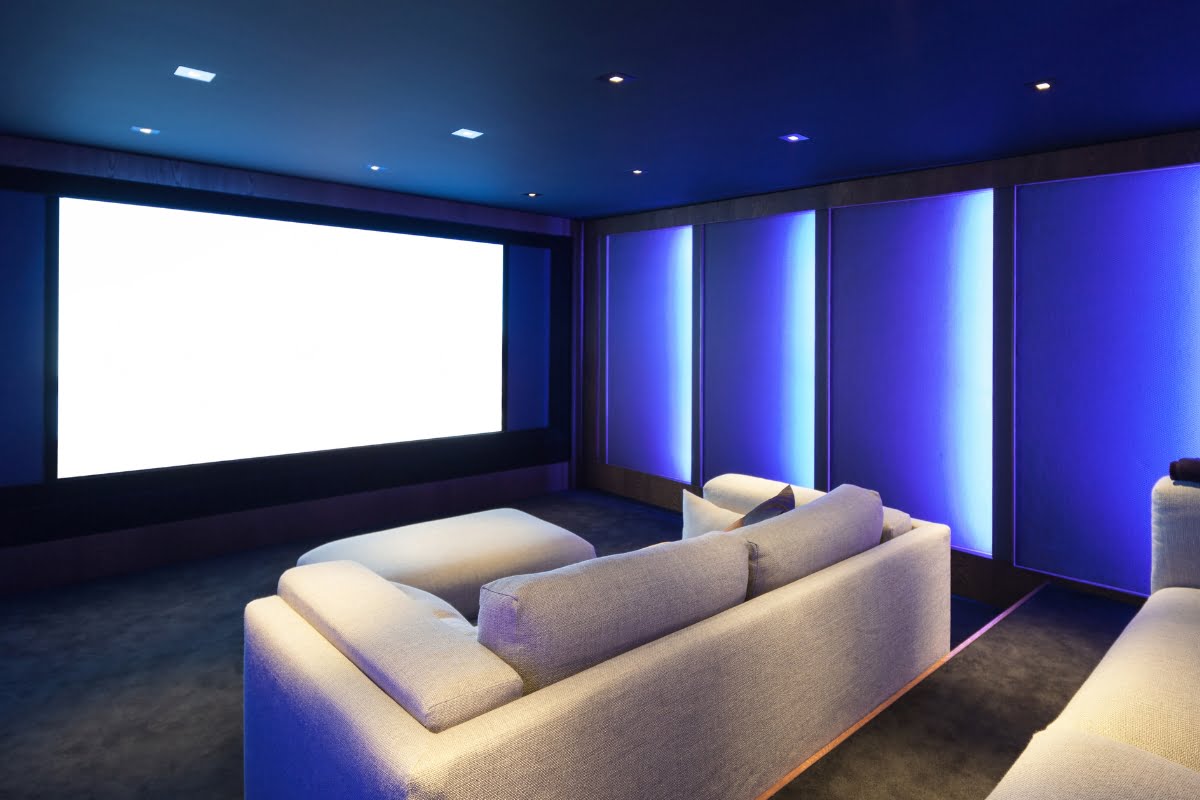 A modern home theater with blue accent lighting, a large projection screen, white sofas arranged for viewing, and integrated home automation systems.