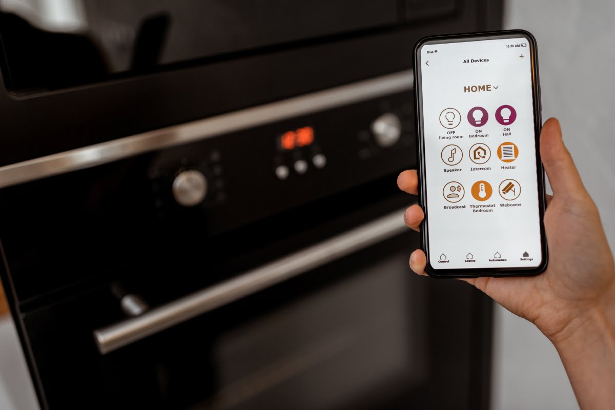 Person holding a smartphone showing a home automation app with various icons, highlighting the seamless integration of home automation systems, in front of a black oven.