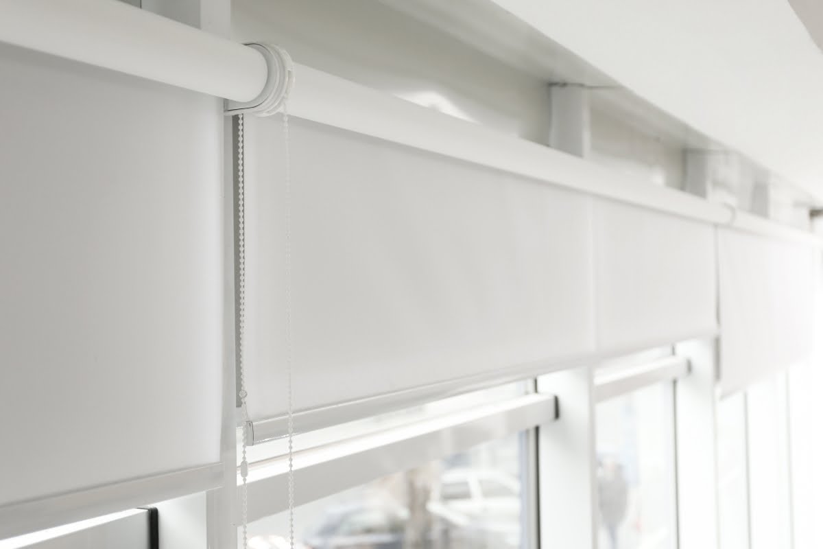 Close-up view of a white roller shade installed on a window, with a pull chain hanging on the left side. The window provides a partial view of cars outside, highlighting how home automation systems can seamlessly integrate into everyday settings.