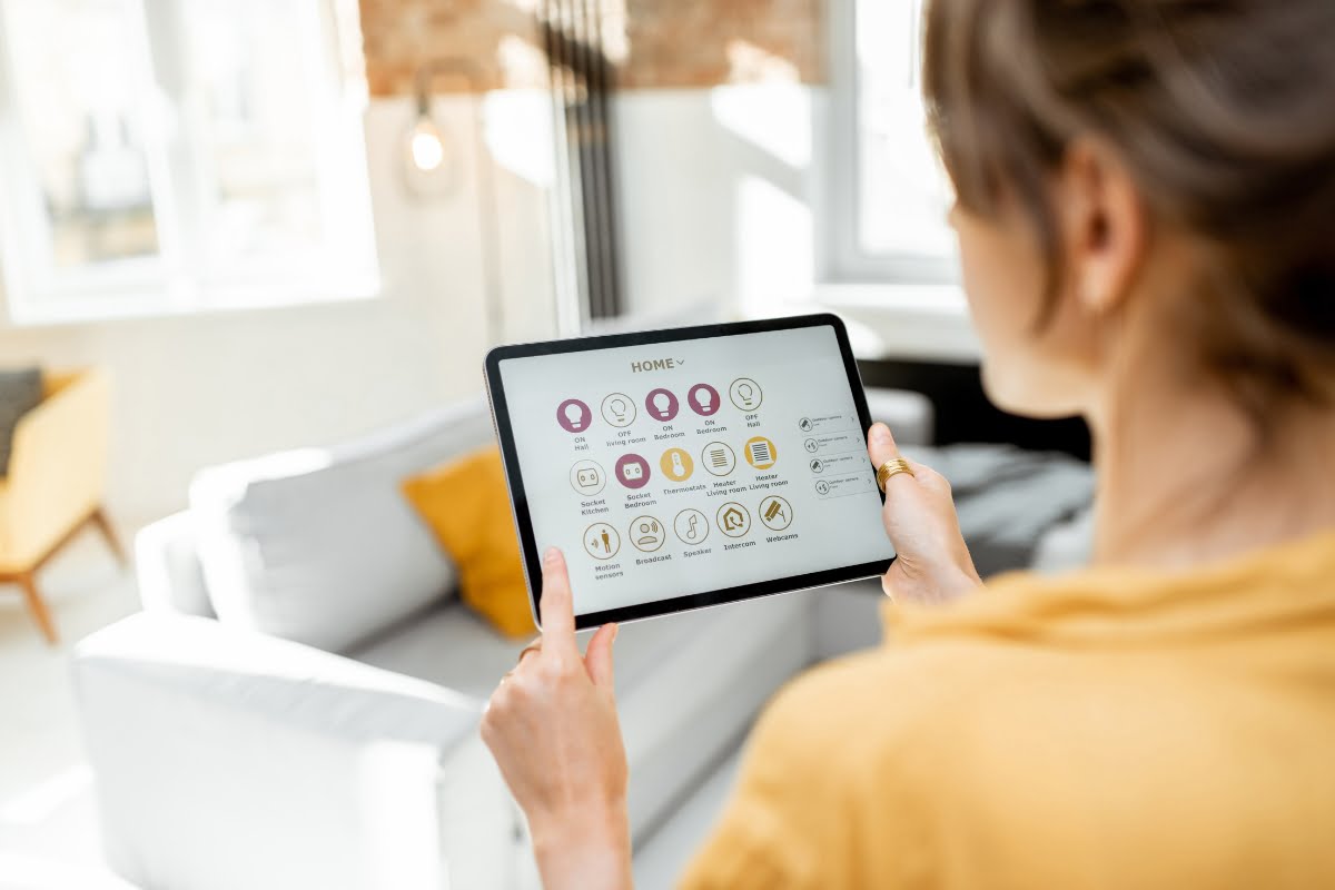 Person using a tablet to control home automation systems while standing in a living room with a white sofa and yellow cushion.