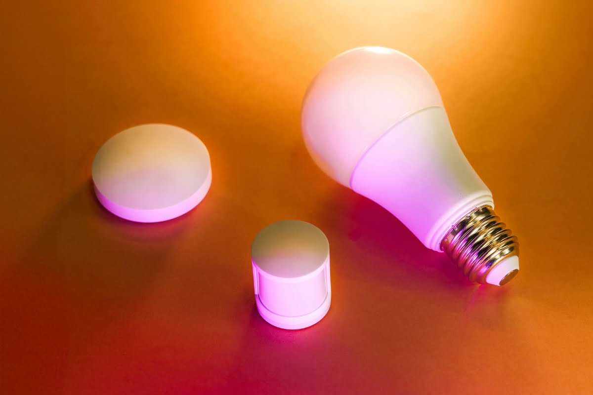 A light bulb and two round, small electronic devices with glowing purple lights, integral to home automation systems, are placed on an orange surface.