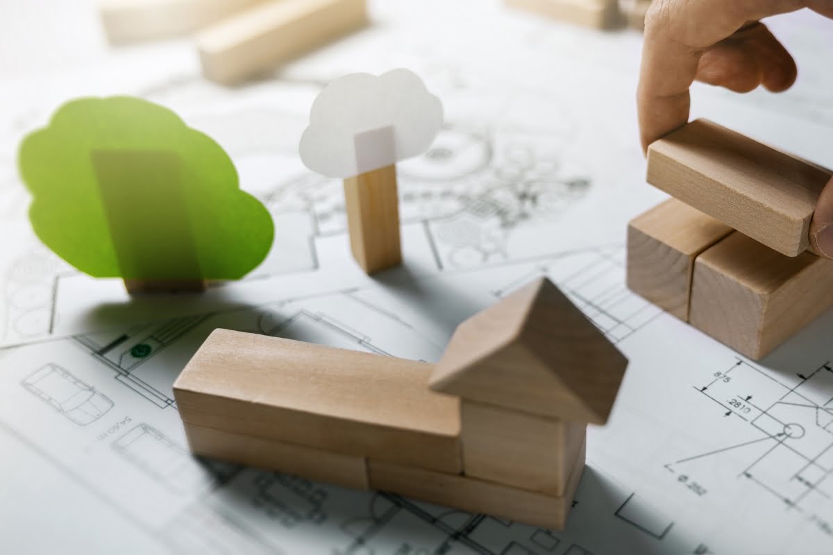 Hand placing wooden blocks on an architectural plan, with green and white wooden trees nearby, highlighting the use of sustainable building materials.