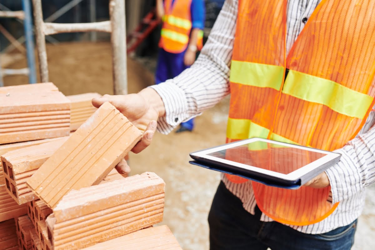 Construction worker in an orange vest holds a tablet in one hand and picks up a brick with the other at a construction site, focusing on sustainable building materials.