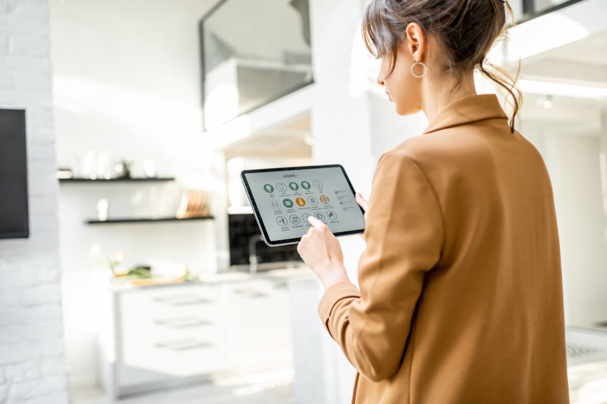 A woman in a brown jacket uses a tablet to control smart home features, standing in a modern kitchen. The tablet screen displays various home automation options, highlighting her savvy home investment.